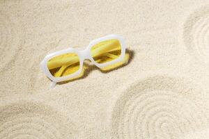 Modern White sunglasses on beach sand background at sunlight with shadow. Summer fashion eyeglasses with yellow glass. Summer vacation, summer rest concept. Minimal style trend flat lay photo