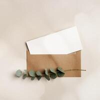 Blank paper card mockup and craft envelope, eucalyptus plant on beige table. White empty letter or invitation, sunlight, beautiful soft shadow. Minimal lifestyle photo. Flat lay, top view, copyspace photo
