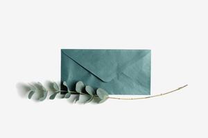 Teal color envelope, blank paper card mock up, branches eucalyptus leaves on white background. Empty colored invitation on table. Minimal modern lifestyle photo. Flat lay, top view, copyspace photo