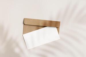 Blank paper card mock up and craft envelope on beige background and palm leaf shadow. White empty invitation on table. Minimal fashion lifestyle photo. Summer Flat lay, top view, copy space. photo
