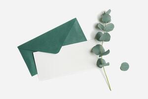 Teal envelope and empty paper card mock up with green plant leaves eucalyptus on white background. White blank letter on table. Minimal design stationery, flat lay template, top view, copy space. photo
