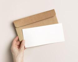 Blank paper card mock up in woman hand, craft envelope on beige background. White empty invitation on table. Female open paper letter. Minimal lifestyle photo. Flat lay, top view, copy space. photo