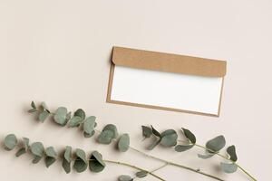 Blank paper card mock up and craft envelope with branches eucalyptus leaves on beige background. White empty invitation on table. Minimal lifestyle photo. Flat lay, top view, copy space. photo