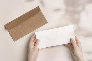 Blank paper card mock up in woman hands, craft envelope on beige background with shadow at sunlight. White empty invitation Female open paper letter. Minimal Flat lay, top view, copy space. photo