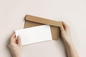 Blank paper card mock up in woman hands, craft envelope on beige background. White empty invitation on table. Female open paper letter. Minimal lifestyle photo. Flat lay, top view, copy space. photo