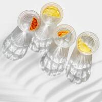 Crystal glasses with citrus slices in water on white background with palm leaf at sunlight, modern, minimalist composition with clear glass cups with water and citrus fruit, as lemon, orange photo