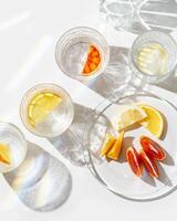 Citrus Infused Water in clear Glasses casting artistic light rainbow reflections and shadows at sunlight. Summer drink concept, minimal style pattern, flavored water with yellow red slices of fruit photo