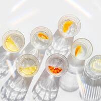 Fruit Infused Water in transparent Glasses with Shadows at sunlight on White background. Aesthetic view summer healthy drink with slices of lemon, orange with rainbow sun glare from glass cups photo