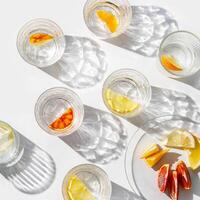 Citrus Infused Water in clear Glasses and plate, artistic light reflections and shadow in natural sunlight. Summer drink concept, minimal pattern, flavored water with yellow red slices of fruit, photo