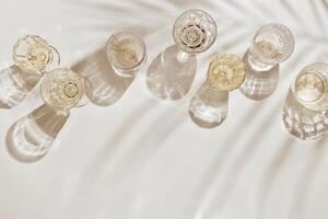 Summer party drinks flat lay, wine glasses with white sparkling wine and palm leaf shadow on light table. Minimal pattern with beautiful wine glasses, above view still life, beige golden neutral color photo