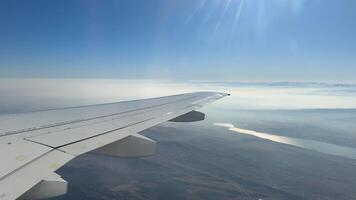 Serene view from an airplane window showcasing the wing with clouds and mountains in the background. The landscape below includes a shimmering lake under a clear sky video