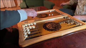 Two adults make moves in backgammon, concentrating on the game. Wooden board with unique design and pieces creates an atmosphere of comfort and strategy video