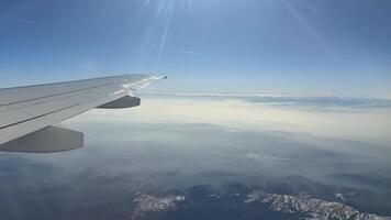 Expansive view from an airplane window featuring the wing, clouds, and distant mountains. A serene landscape unfolds below under a clear blue sky video