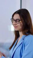 Brunette business lady is calling somebody on the phone. Man at backdrop using laptop standing at the window. Blurred background. Vertical video