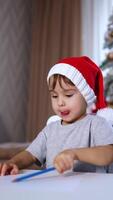 Beautiful baby boy in red cap sitting at desk. Toddler drawing with a blue pencil. Christmas tree at backdrop in blur. Vertical video