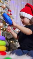 Toddler boy in black t-shirt playing with toy pyramid. Kid wearing Santa cap assembling toy near the Christmas tree. Low angle view. Vertical video