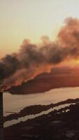 Massive air pollution near the river at darkness. Huge puffs of harmful smoke exhaust into the air against setting sun. Chimneys with dark fumes from manufacture. Aerial view. Vertical video
