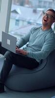 Comfortable work conditions in the modern office. Happy young man sitting in bean bag chair typing on his laptop at looking at window. High angle view. Vertical video