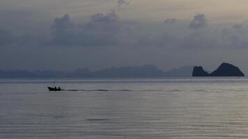 A silhouette of a lone fisherman in a small wooden boat moves across a tranquil sea at dusk, leaving a gentle wake behind. The distant islands and dramatic cloudscape create a serene and atmospheric video
