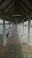 A long, covered pier stretches over the water, creating a stunning perspective effect with its symmetrical roof structure and supporting pillars. The converging lines of the walkway lead toward a video