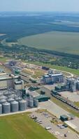 Large industrial territory among beautiful nature. Modern industry for keeping and processing crop. Agricultural complex with grain elevators on field in summer. Aerial view. Vertical video