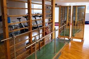 A rehabilitation and physiotherapy gym equipped with parallel bars, wall bars, and mirrors for physical therapy exercises. The space is designed to aid mobility training and recovery. Various exercise photo