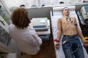 A female doctor performs an ECG on a young man in a hospital room, carefully monitoring his heart health during a routine checkup. photo