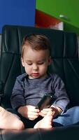 Calm Caucasian child sitting in a comfortable office holding remote control. Lovely boy looks away and smiles adorably to approaching camera. Low angle view close up. Vertical video