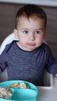 Baby boy sits in a high chair holding it on two sides. Little kid is fed from spoon while he looks up attentively. Low angle view. Vertical video