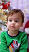 Thoughtful baby boy sitting in front of camera. Lovely toddler near the Christmas tree at home. Blurred backdrop. Vertical video