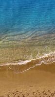 Waves of the sea splash on the sandy beach. Afro-American man stands in the water. Multiple footsteps are on the sand. Top view. Vertical video