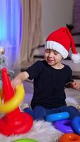 Peaceful toddler in Santa cap playing at Christmas tree. Little boy collecting colorful pyramid on the floor. Vertical video