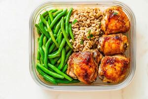 Flat lay of a meal prep container featuring baked chicken thighs, brown rice, and fresh green beans, arranged in a glass container on a white background photo