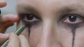 Makeup artist's hands carefully applying dark eyeshadow around model's eyes, creating a dramatic look with black tears for a goth rock or Halloween event video