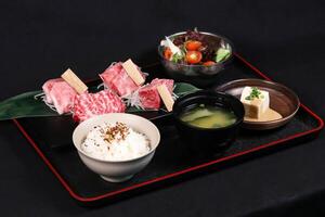 Wagyu Beef Yakiniku Bento Set on a black tray include raw Wagyu beef slices, rice, miso soup, tofu, and salad isolated on dark background side view of Japanese cuisine photo