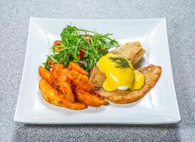 Fried Fish Benedict include fish fillet with a poached egg and hollandaise sauce, alongside salad and wedges served on a plate side view of hong kong food on a textured stone background photo