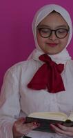 Close-up of a Muslim woman wearing a white headscarf, reading the Quran with Arabic script. She is deeply focused on the sacred text, reflecting faith and devotion. Isolated against a pink background. video