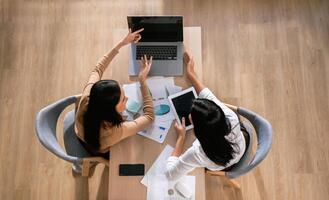 Two women are sitting at a table with laptops and papers in front of them photo