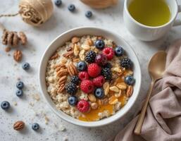On the light gray surface there is a plate with oatmeal, decorated with fresh berries ,raspberries, blueberries, blackberries, and nuts, sprinkled with honey. Next to a cup of green tea, top view photo