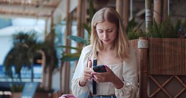 Blonde girl summer clothes stands tropical cafe scrolling through her phone. Embracing technology, she explores technology through device. Technology connects, enhances, transforms daily experiences. video