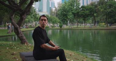 Sad lonely man with smartphone in his hand is waiting for a girlfriend on date, but she did not come. Depressed man, left alone on bench in city park near lake, suffers from breakup with a girlfriend. video
