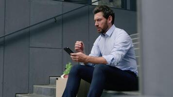 Serious fired unemployed Caucasian middle-aged businessman using smartphone searching work vacancy online thinking business man male guy in city with mobile phone with dismissal box dismiss lost job video