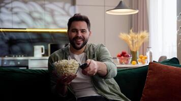 Happy glad Caucasian laughing business man male guy homeowner spectator relaxing at home sofa holding remote TV controller choosing film laugh comedy watching television program eating popcorn indoor video