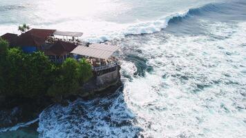 Scenic coastline with cafe on rock and ocean waves on Nusa Ceningan, Bali. Drone view video