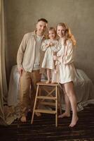 Family portrait captured in a cozy indoor setting featuring a smiling father, mother, and daughter standing on a stool against a neutral background photo