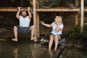 Friends enjoying a summer day by the water, one hanging from a platform and the other seated on a log, both laughing joyfully in a natural setting photo
