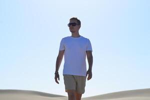 A stylish portrait of a young man walking along a sand dune through barkhan in Maspalomas, Gran Canaria on a hot sunny day. Desert travel touristic concept photo