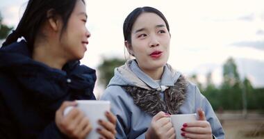 Handheld selective focus shot, Two Asian female are sitting in a garden holding a coffee cup and enjoying the view in front of them on a cold morning. Winter activity. video