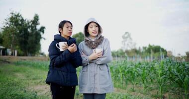 Two Asian women standing talking in a garden holding cups of coffee and enjoying the view in front of them on a cold morning. Winter activities. video