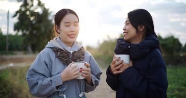 Handheld shot, Two Asian women standing talking in a garden holding cups of coffee and enjoying the view in front of them on a cold morning. Winter activities. video